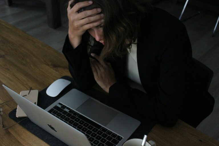A woman sits at a laptop with her head in her hand, looking mentally drained and stressed