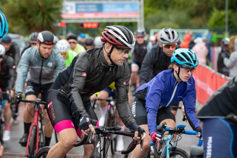 Richard Davison in a black rain jacket and Cycling Science bib shorts, riding at the front of a bunched group at the wet start of a mass-participation road event, passing under the timing gantry.