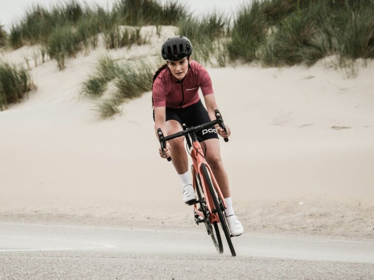 A woman in a maroon jersey riding a road bike at speed along a coastal road beside sand dunes.